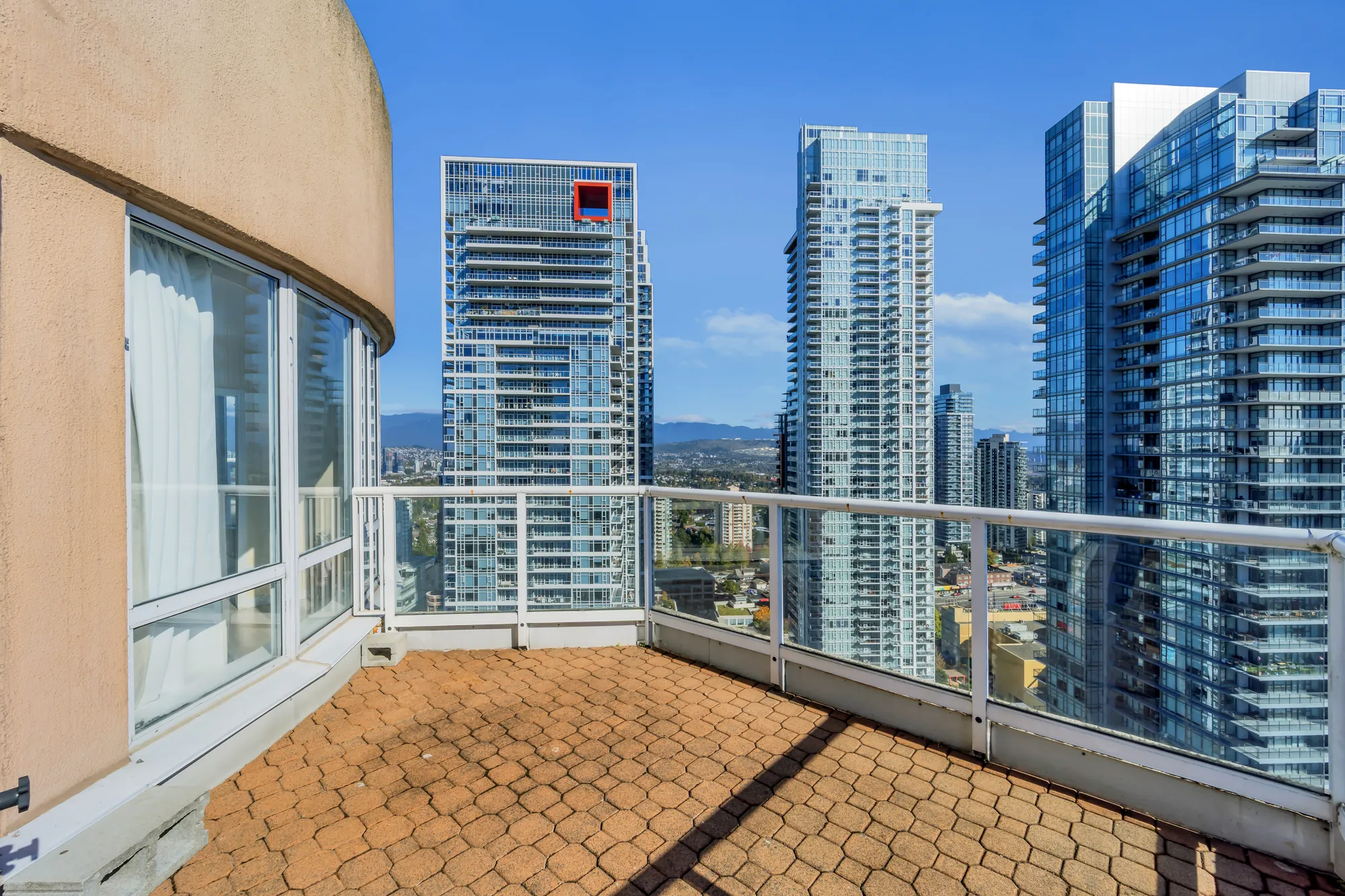 A balcony with a paver floor and a glass railing, the curved wall of the building at the left, towers of Metrotown beyond