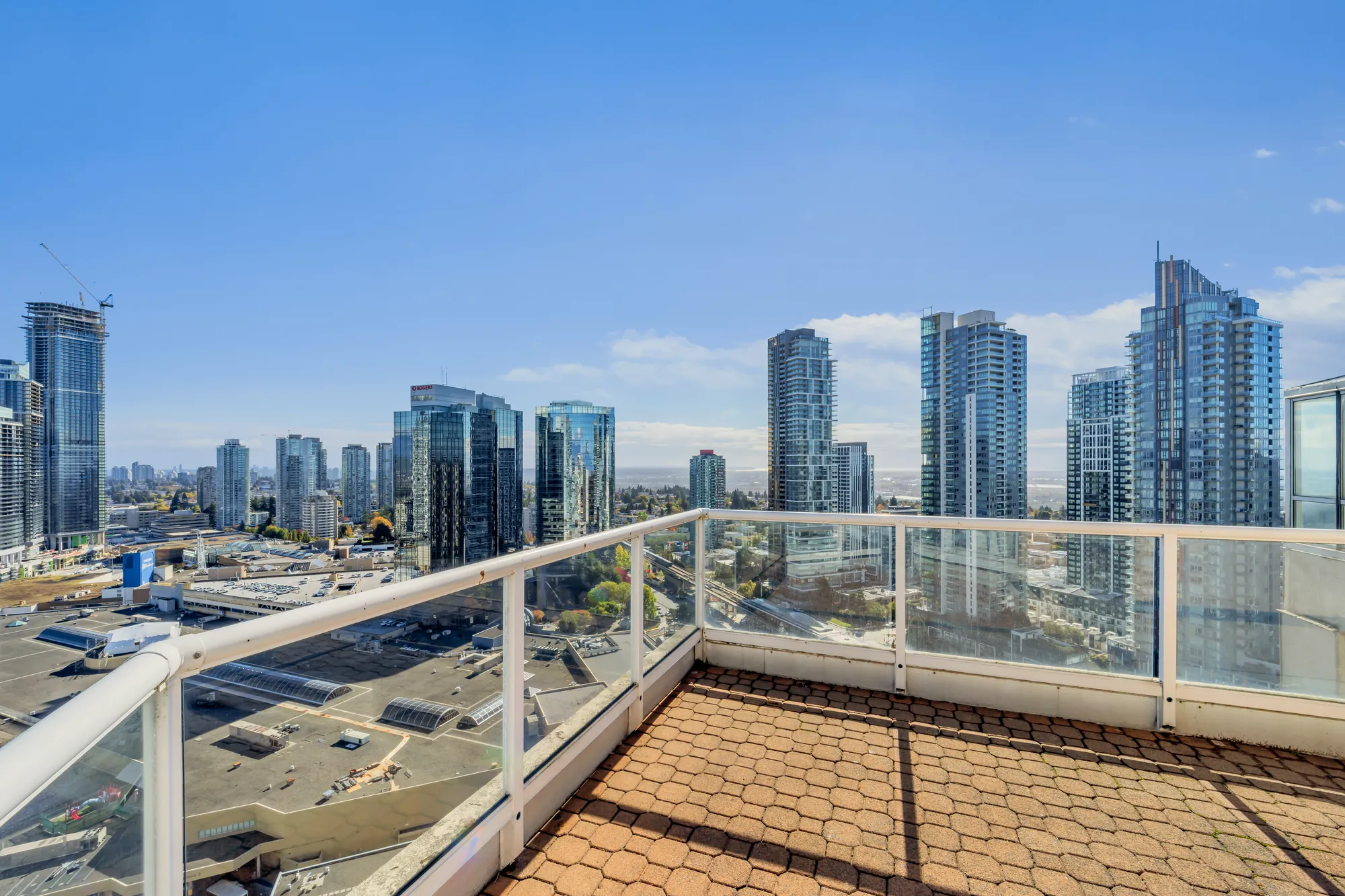 The corner of a top-floor balcony at 2804-6220 McKay Avenue: a paver floor and a glass railing, the towers of Metrotown and the city to the horizon under a blue sky