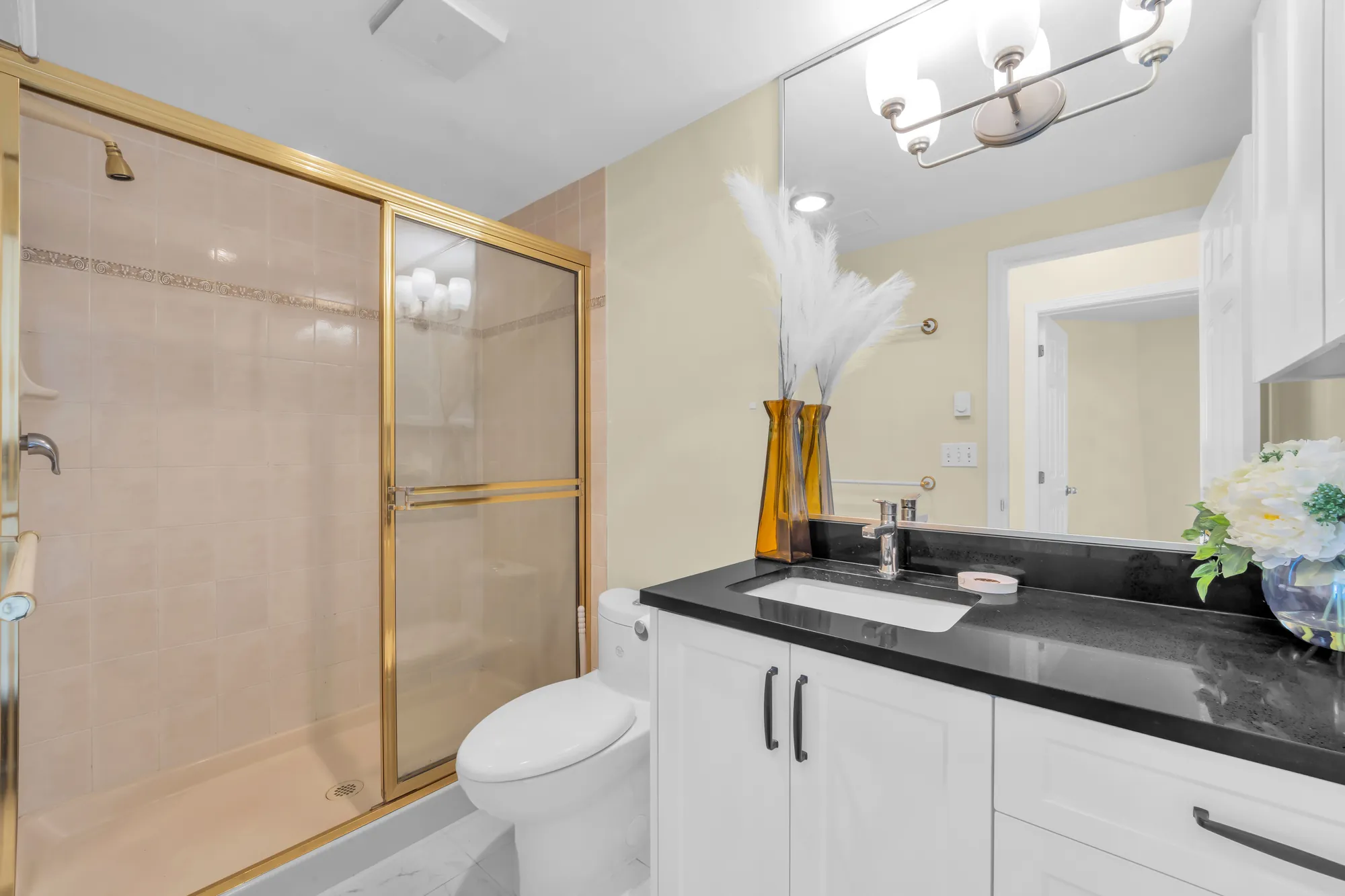 A bathroom: a shower behind gold-framed glass doors, a white vanity with a black counter, a wide mirror and a light bar