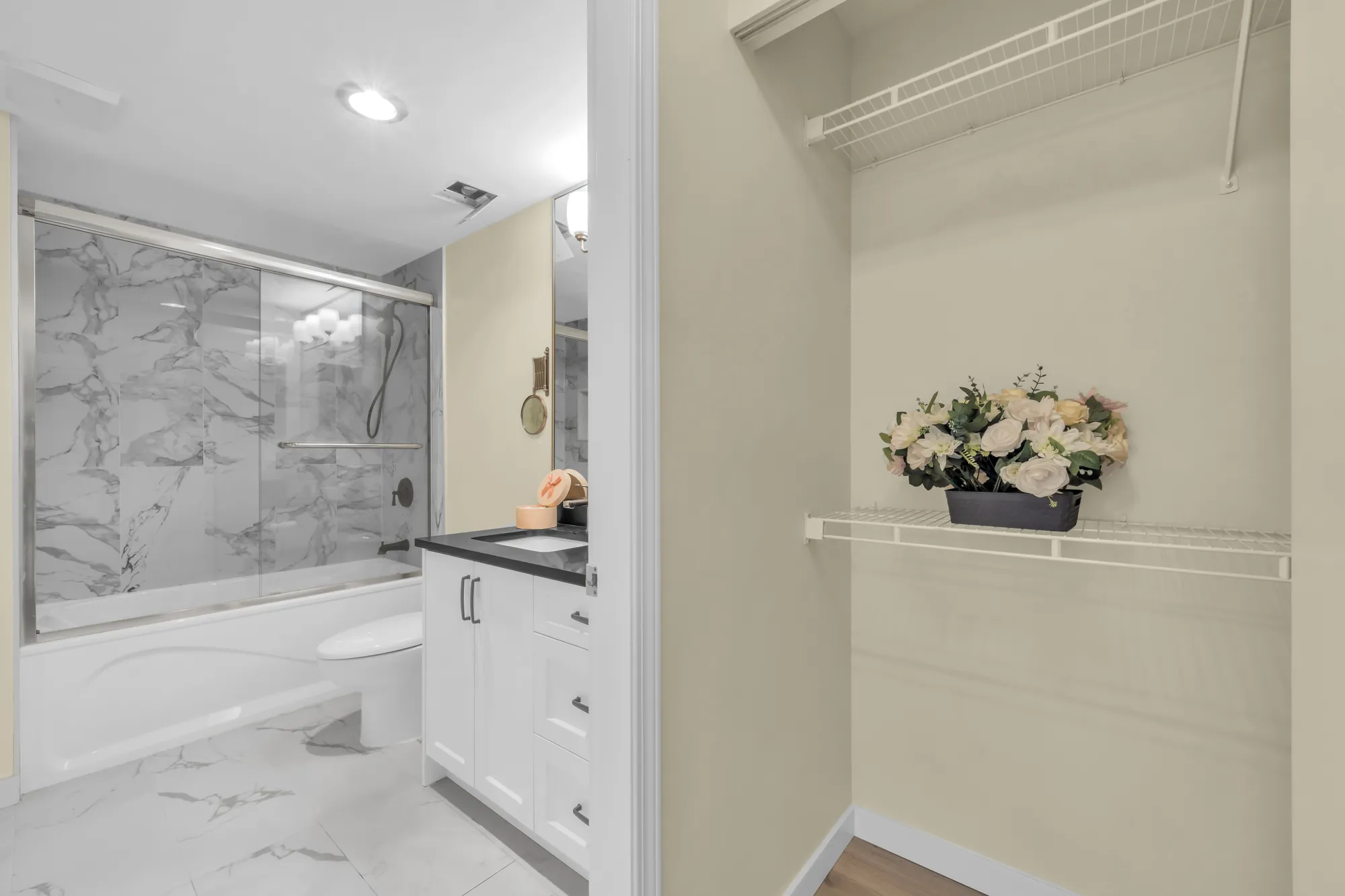 A bathroom with a tub and shower in marble-look tile, a white vanity, and a closet with wire shelves beside it