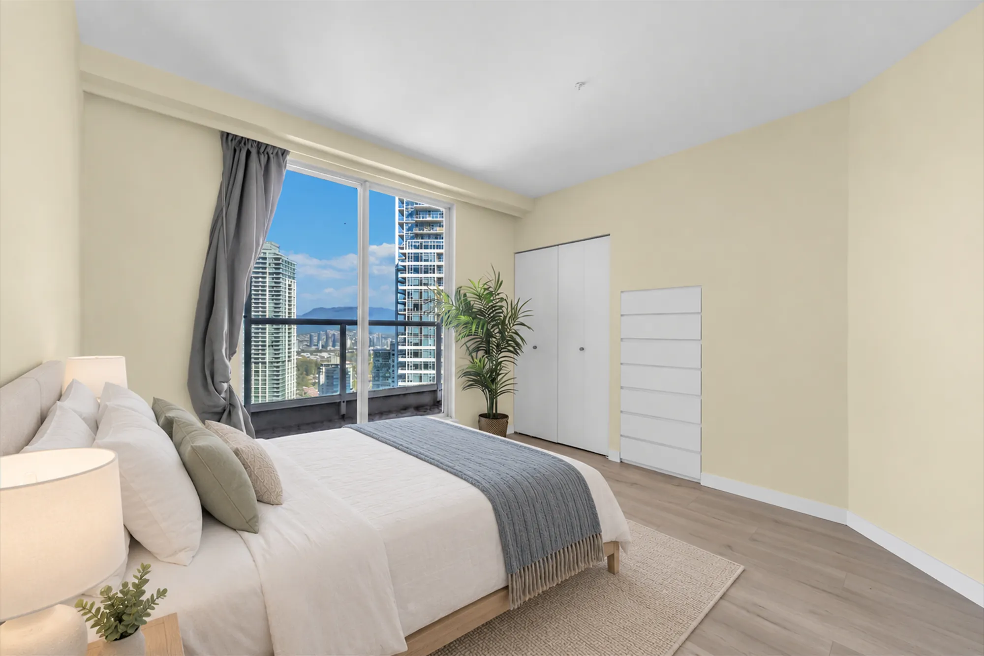 A bedroom with a sliding glass door to a balcony, virtually staged: an upholstered bed with a grey throw, a night table and lamp, a rug, a palm by the window, closets with white doors