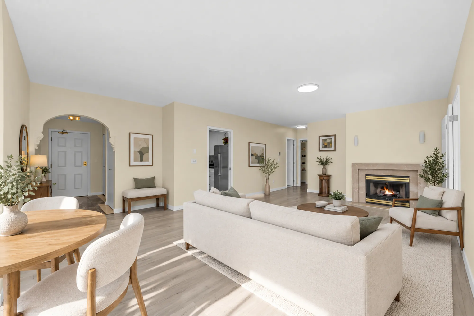 The living room, virtually staged: a cream sofa and armchairs around a round coffee table on a rug, a round wood dining table with white chairs in the foreground, the gas fireplace in a stone surround, an arched doorway to the entry