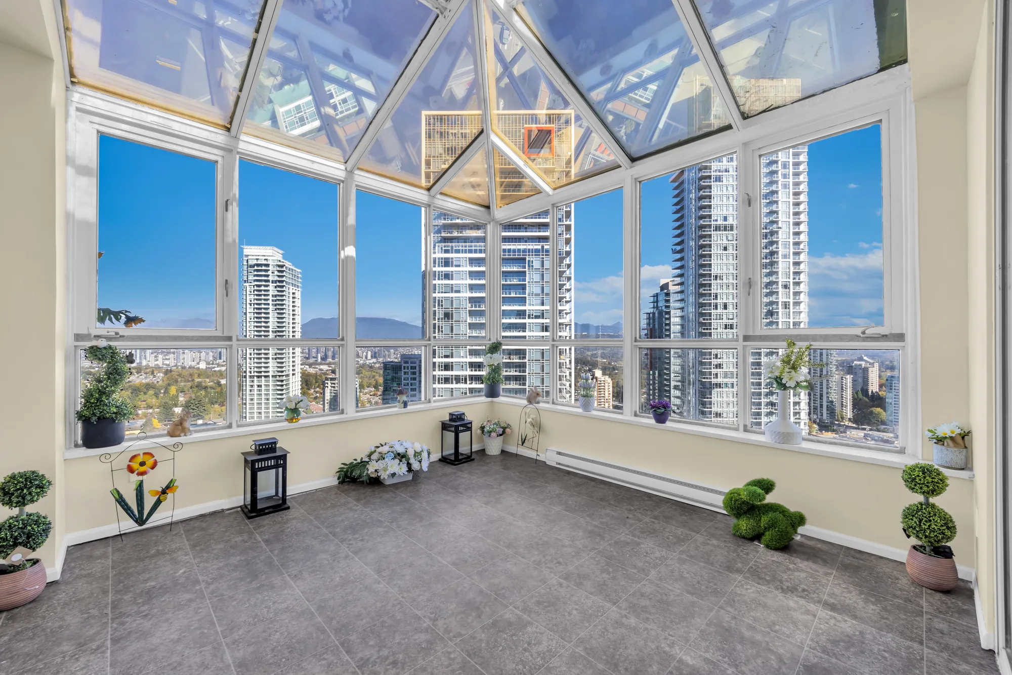 The sunroom: glass on three sides and a glass roof on white frames, a grey tile floor, towers and the mountains beyond, ornaments and potted plants along the sills