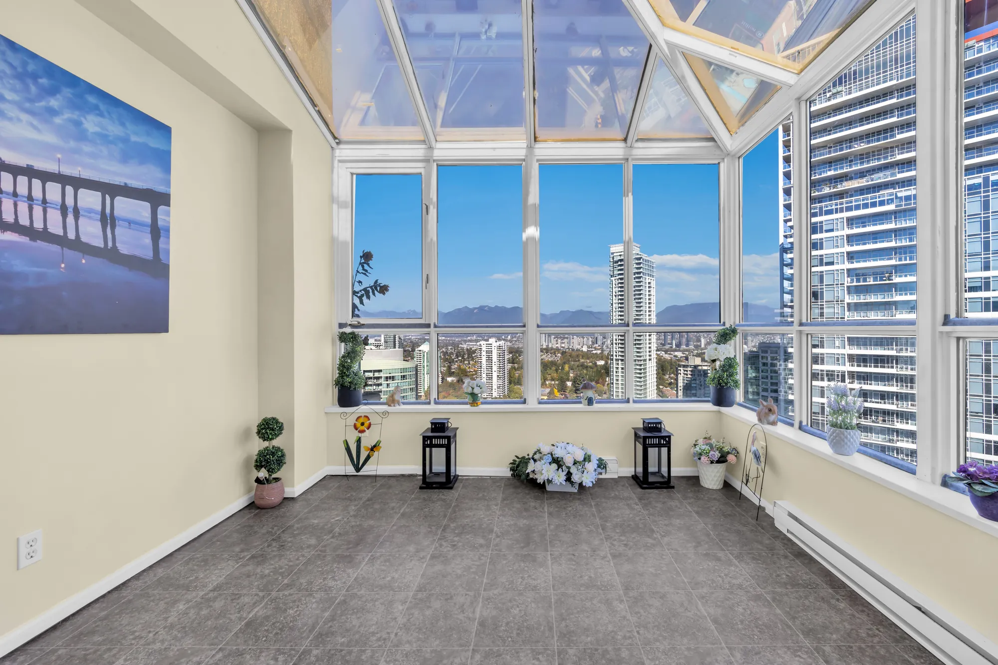 The sunroom from its doorway: the glass roof, windows on two sides onto the towers of Metrotown and the mountains, a framed print on the wall